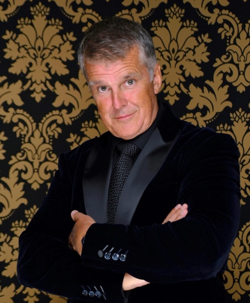 Dominic Pearce wearing a dark suit and tie, standing with his arms folded in front of a decorative black and gold background.